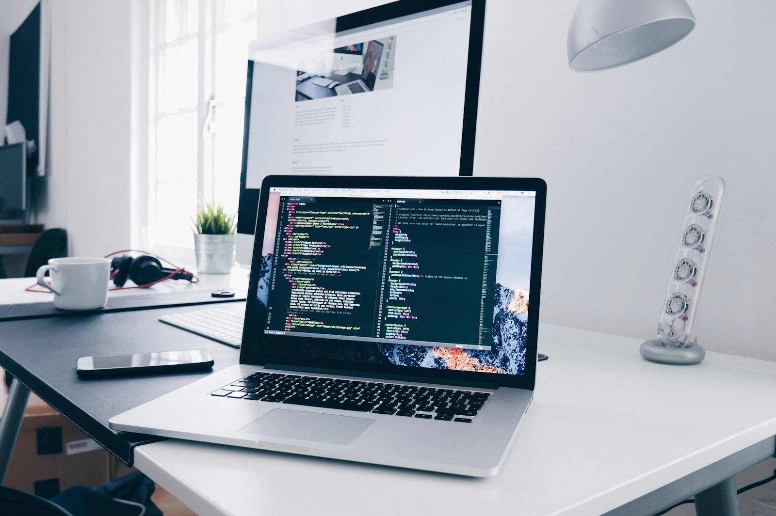 A laptop displaying code on a bright desk beside a large monitor.